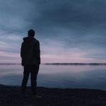 A solitary silhouette of a man in a jacket gazing at a lake during a peaceful sunset, creating a serene atmosphere.