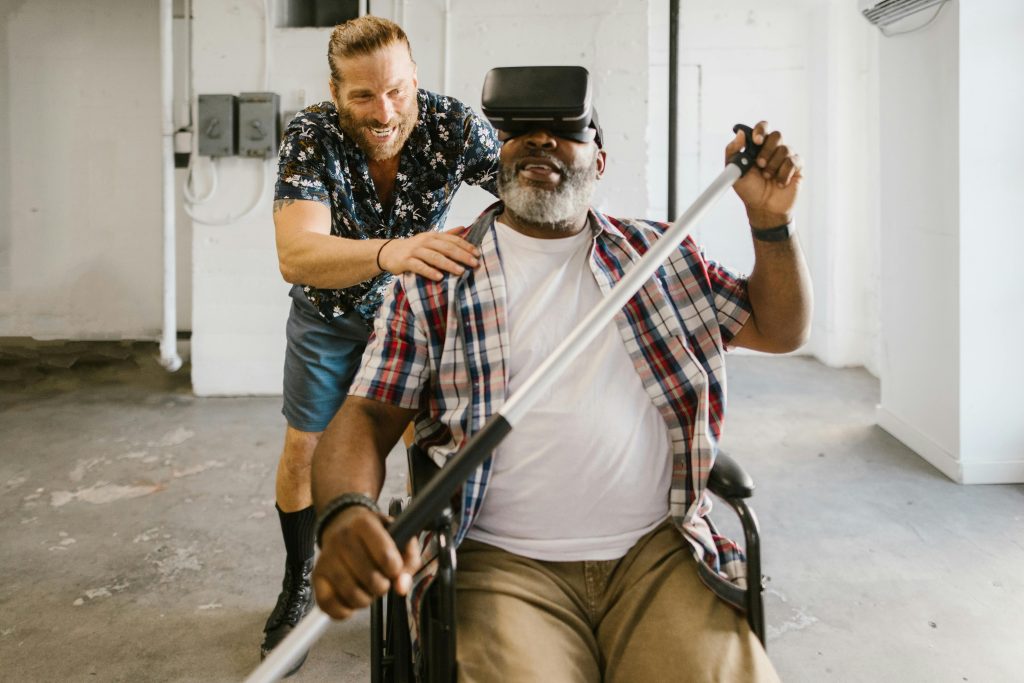 Man assists wheelchair user engaged in rehabilitation exercise with VR set indoors.